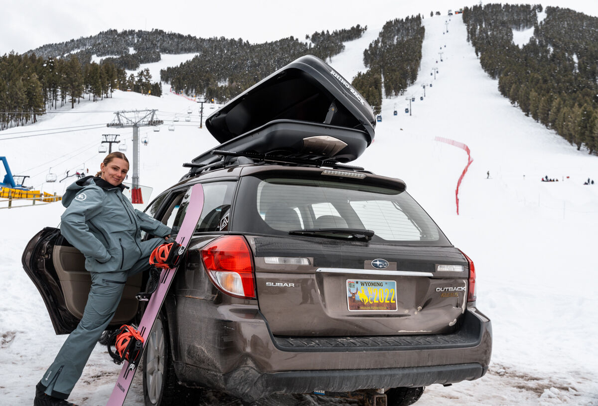 A blond Women snowboard model standing Next to car at base of ski slope ready to snowboard