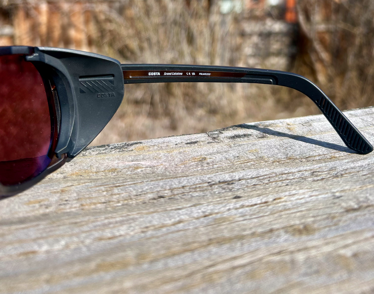 Costa Del Mar high-performance sunglasses with blue mirror lenses, showcased at the summit of Teton Pass, Wyoming. The background features the iconic rolling mountains and steep terrain where the gear was Teton Tested for clarity and glare reduction.