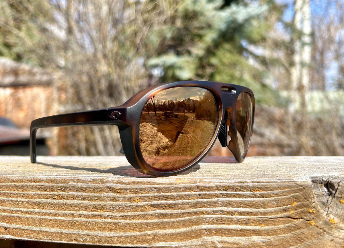 A close-up shot of Costa Del Mar sunglasses featuring polarized 580 lenses, resting against a backdrop of the rugged Teton Pass mountain range in Wyoming. The high-altitude scenery shows a vast valley and snow-dusted peaks under a clear blue sky, highlighting the eyewear's design for high-performance outdoor adventure.
