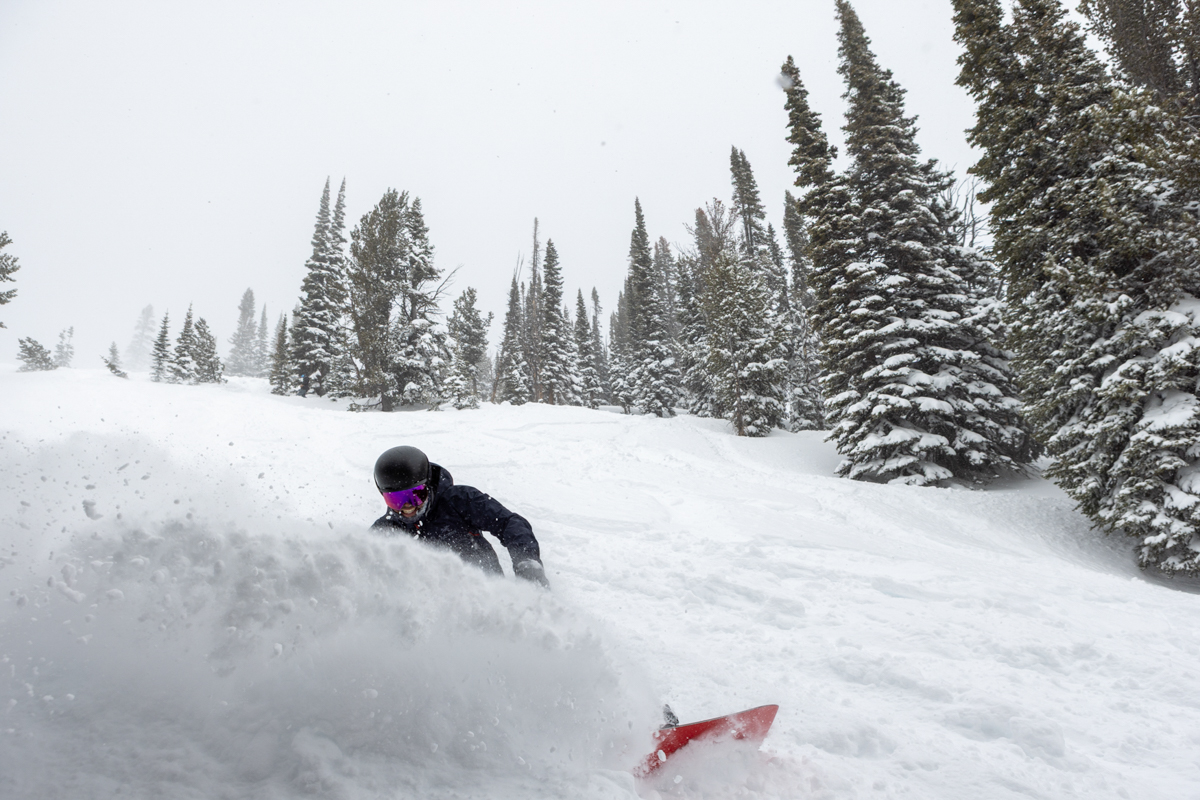 Alt Text: Snowboarder slashing a powder turn near trees on the Korua Transition Finder snowboard.