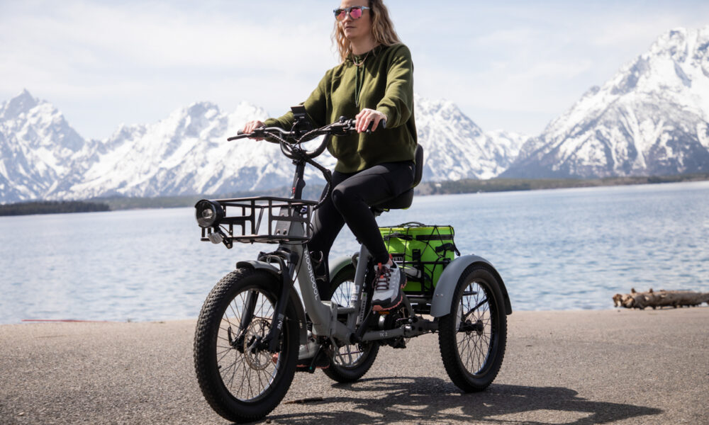 Woman in a green jacket riding the Mooncool TK2 trike toward the camera with mountain scenery