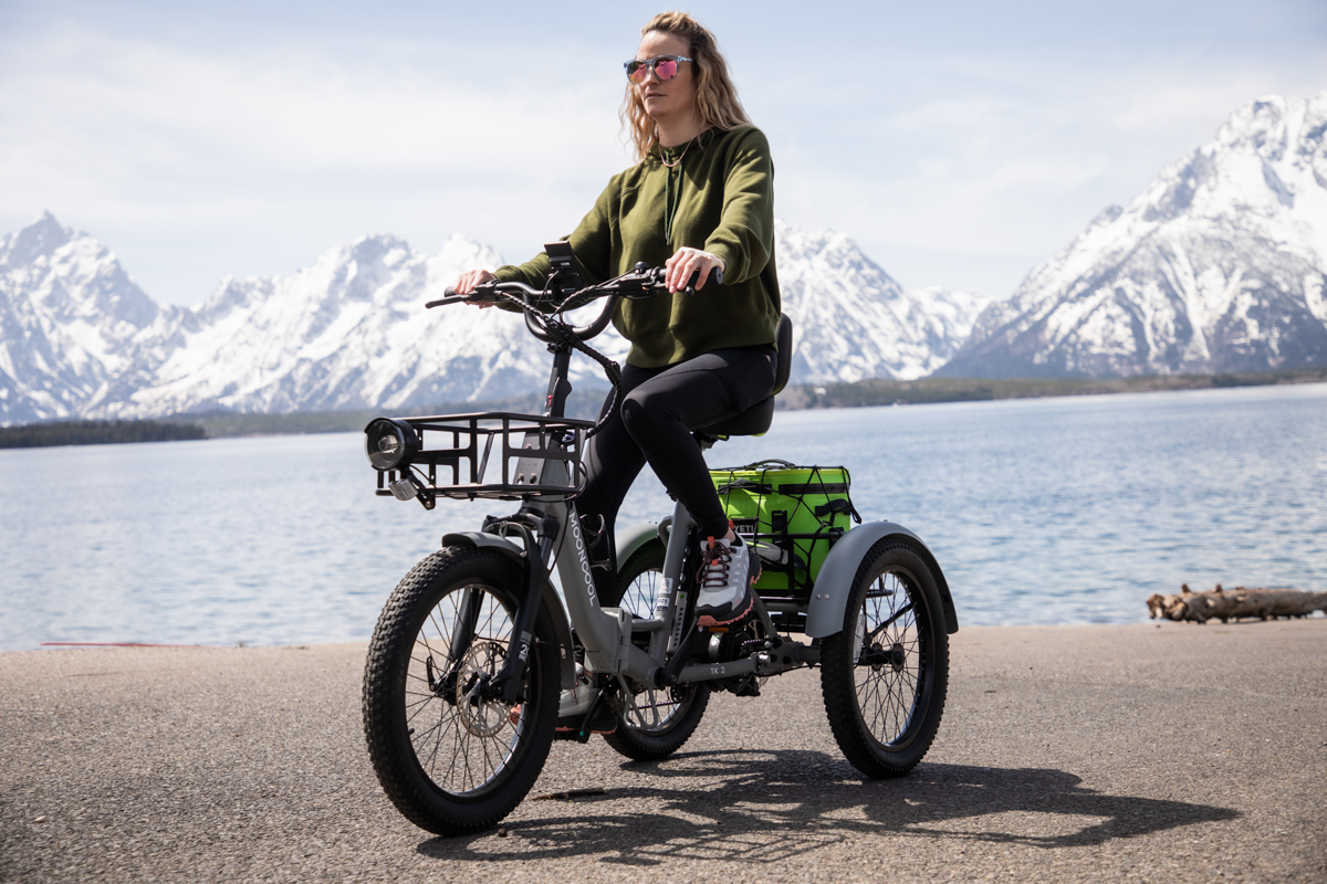 Woman in a green jacket riding the Mooncool TK2 trike toward the camera with mountain scenery