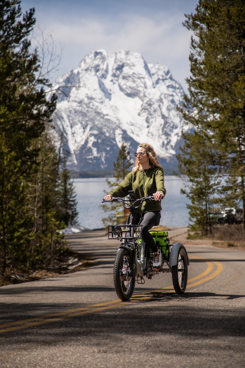 Woman riding a white Mooncool TK2 electric trike on a paved road with the Teton mountains in the background.