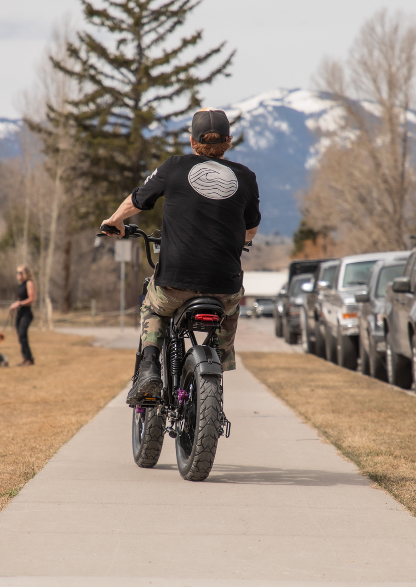 Rear view of a rider cruising the Talon T87 E-Bike on a paved pathway near downtown Jackson, WY, showcasing its versatility from dirt trails to city streets.